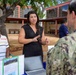 An outdoor informational or outreach event is taking place in what appears to be a courtyard or campus setting. A woman in a black dress is actively presenting or explaining something to a person in U.S. Navy digital camouflage (NWU) uniform, while another woman in a striped shirt stands nearby. A table with a laptop, printed materials, and a green folder is visible in the foreground.