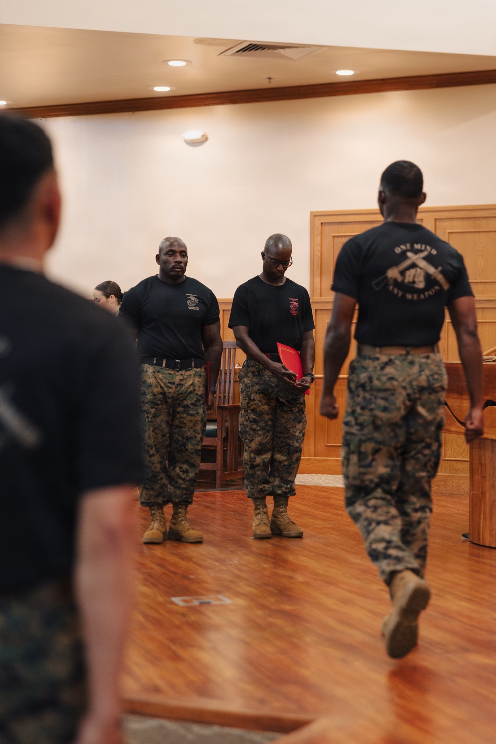 The image shows a group of U.S. Marines in a formal indoor setting, likely a chapel or ceremonial room based on the wooden paneling and pew-like furniture. Several Marines in black PT shirts and MARPAT camouflage trousers are gathered, with one holding a red folder appearing to read or present. The back of one Marine's shirt reads 'ONE MIND ANY WEAPON,' a phrase associated with Marine Corps Martial Arts Program (MCMAP).