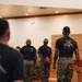 The image shows a group of U.S. Marines in a formal indoor setting, likely a chapel or ceremonial room based on the wooden paneling and pew-like furniture. Several Marines in black PT shirts and MARPAT camouflage trousers are gathered, with one holding a red folder appearing to read or present. The back of one Marine's shirt reads 'ONE MIND ANY WEAPON,' a phrase associated with Marine Corps Martial Arts Program (MCMAP).