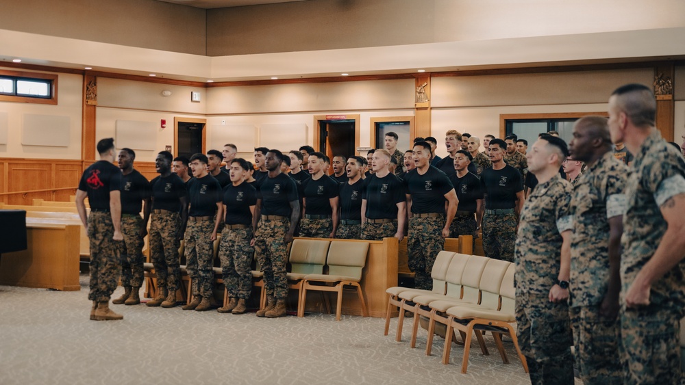 A large group of U.S. Marines in black PT shirts and camouflage trousers are standing in formation inside what appears to be a chapel or auditorium, singing or reciting in unison. Several senior Marines in full camouflage uniforms observe from the right side of the frame. The setting suggests a formal military ceremony, motivational event, or unit gathering.