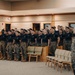 A large group of U.S. Marines in black PT shirts and camouflage trousers are standing in formation inside what appears to be a chapel or auditorium, singing or reciting in unison. Several senior Marines in full camouflage uniforms observe from the right side of the frame. The setting suggests a formal military ceremony, motivational event, or unit gathering.