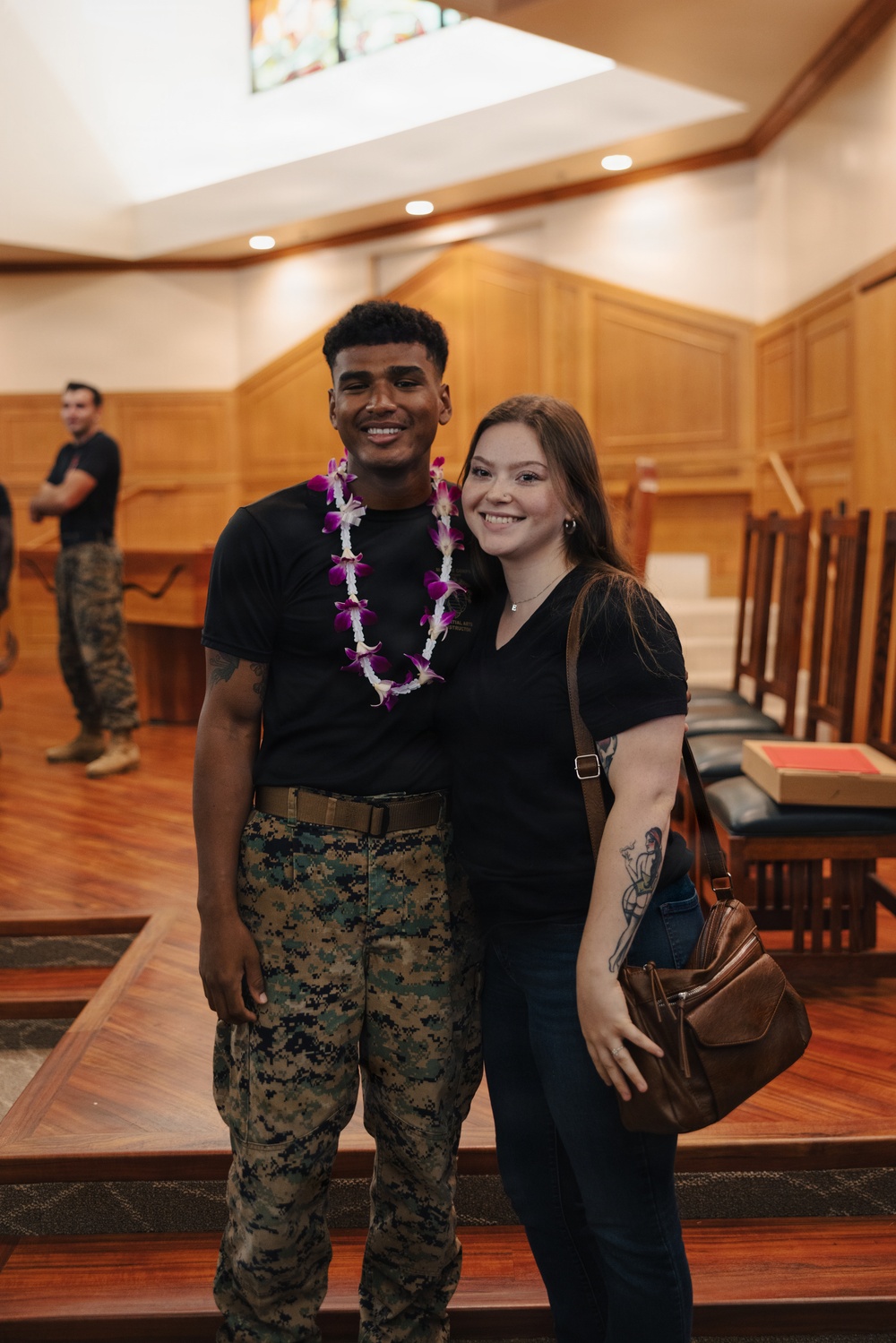 Two individuals pose together smiling inside what appears to be a military chapel with warm wood paneling. The man on the left is wearing U.S. Marine Corps MARPAT camouflage trousers, a black t-shirt, and a purple and white floral lei. A third person in military camouflage pants is visible in the background.