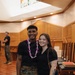Two individuals pose together smiling inside what appears to be a military chapel with warm wood paneling. The man on the left is wearing U.S. Marine Corps MARPAT camouflage trousers, a black t-shirt, and a purple and white floral lei. A third person in military camouflage pants is visible in the background.