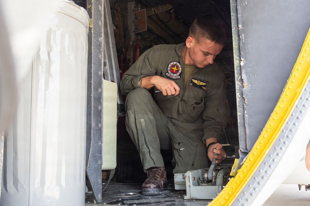 A military personnel member in an olive green flight suit is crouched inside what appears to be a military aircraft, performing maintenance or inspection work on a mechanical component near the floor. The individual wears aviator wings and a squadron patch on their uniform, indicating they are likely a naval or marine aviator or aircrew member. The image shows the interior doorway/hatch area of the aircraft with visible riveted metal panels and a yellow-bordered door frame.