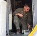 A military personnel member in an olive green flight suit is crouched inside what appears to be a military aircraft, performing maintenance or inspection work on a mechanical component near the floor. The individual wears aviator wings and a squadron patch on their uniform, indicating they are likely a naval or marine aviator or aircrew member. The image shows the interior doorway/hatch area of the aircraft with visible riveted metal panels and a yellow-bordered door frame.