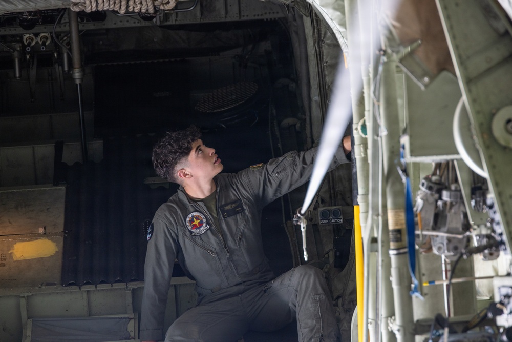 A military personnel member in olive/gray flight suit is inspecting or working on the interior mechanical components of a military aircraft, likely a C-130 Hercules or similar transport aircraft. The individual is looking upward while reaching toward overhead equipment near what appears to be a jump door or cargo ramp area. The setting is the interior of a military aircraft with visible hydraulic lines, cables, and structural components.