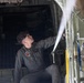 A military personnel member in olive/gray flight suit is inspecting or working on the interior mechanical components of a military aircraft, likely a C-130 Hercules or similar transport aircraft. The individual is looking upward while reaching toward overhead equipment near what appears to be a jump door or cargo ramp area. The setting is the interior of a military aircraft with visible hydraulic lines, cables, and structural components.