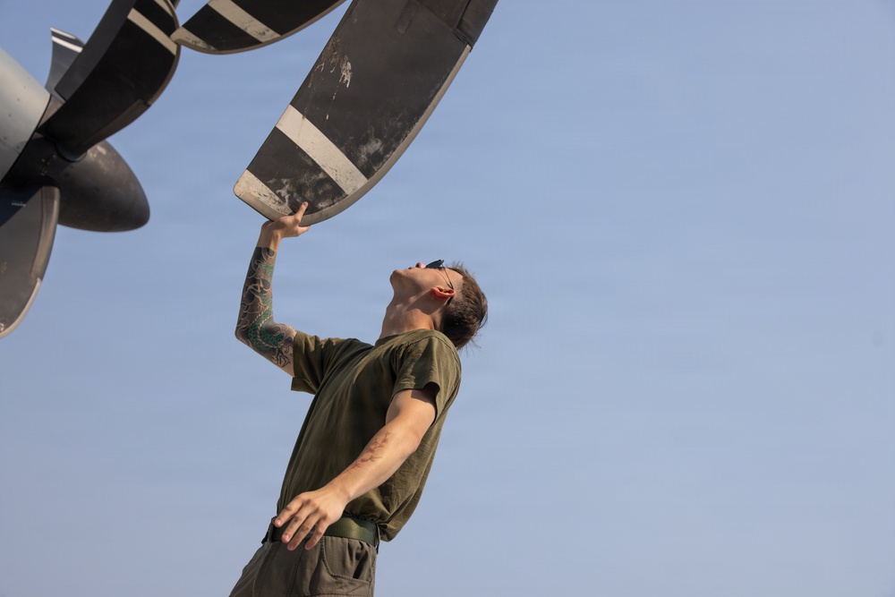 A military or aviation maintenance technician is inspecting a large aircraft propeller blade from below, looking upward while gripping the blade with one hand. The individual is wearing an olive green military-style t-shirt and has visible tattoo sleeve on one arm. The photo is taken from a low angle against a clear blue sky, emphasizing the scale of the propeller.