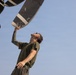 A military or aviation maintenance technician is inspecting a large aircraft propeller blade from below, looking upward while gripping the blade with one hand. The individual is wearing an olive green military-style t-shirt and has visible tattoo sleeve on one arm. The photo is taken from a low angle against a clear blue sky, emphasizing the scale of the propeller.