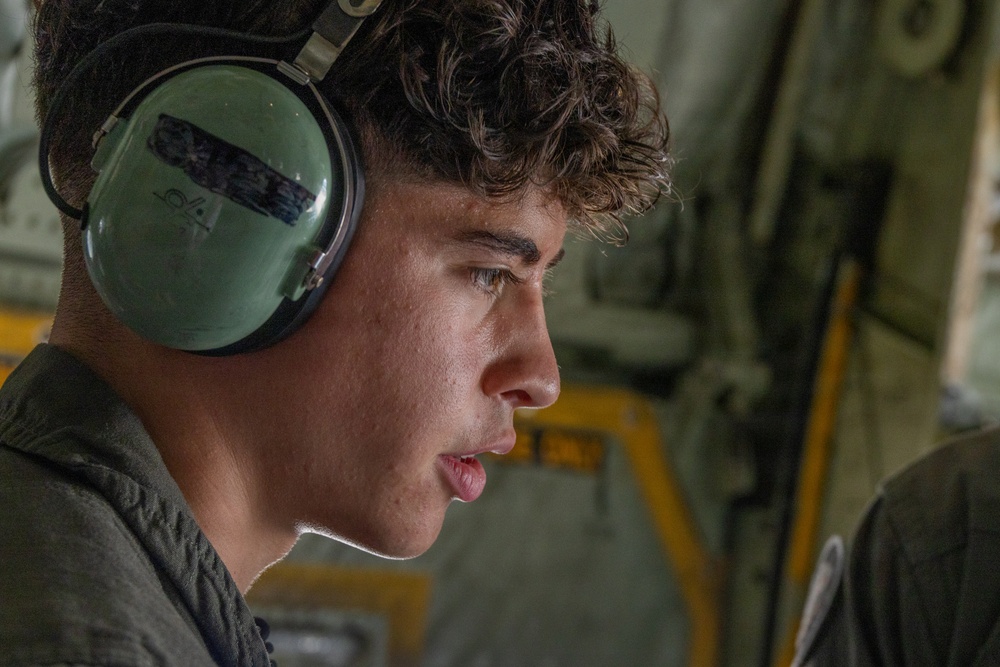 A young male individual wearing military-style olive green hearing protection headset is photographed in close profile view inside what appears to be a military aircraft. The subject appears to be speaking or communicating, possibly via the headset. The background reveals the interior of a military transport aircraft with visible yellow safety markings and olive drab structural elements.