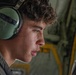 A young male individual wearing military-style olive green hearing protection headset is photographed in close profile view inside what appears to be a military aircraft. The subject appears to be speaking or communicating, possibly via the headset. The background reveals the interior of a military transport aircraft with visible yellow safety markings and olive drab structural elements.