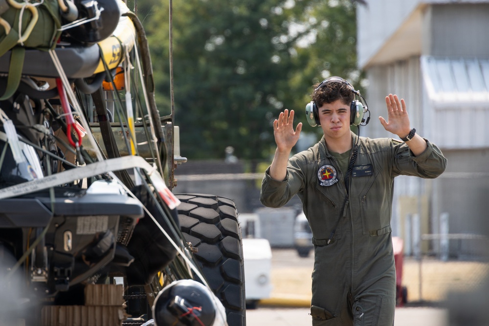 A young military personnel member wearing an olive green flight suit and aviation headset is using hand signals, likely directing ground vehicle movement on a military flight line or tarmac. A large military vehicle or ground support equipment is visible in the foreground left, loaded with various gear and equipment. The setting appears to be a military airfield or base.
