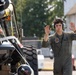 A young military personnel member wearing an olive green flight suit and aviation headset is using hand signals, likely directing ground vehicle movement on a military flight line or tarmac. A large military vehicle or ground support equipment is visible in the foreground left, loaded with various gear and equipment. The setting appears to be a military airfield or base.