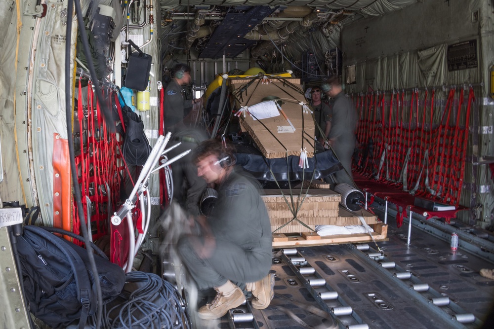 The image shows the interior cargo hold of a military transport aircraft, likely a C-130 Hercules, with crew members loading or securing cargo on pallets. Several personnel wearing flight suits and headsets are visible working around strapped-down cardboard boxes and equipment on a roller-equipped cargo floor. The image has some motion blur on the foreground figure, suggesting movement during the exposure.