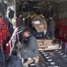 The image shows the interior cargo hold of a military transport aircraft, likely a C-130 Hercules, with crew members loading or securing cargo on pallets. Several personnel wearing flight suits and headsets are visible working around strapped-down cardboard boxes and equipment on a roller-equipped cargo floor. The image has some motion blur on the foreground figure, suggesting movement during the exposure.