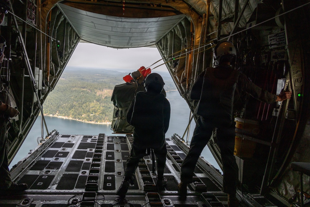 The image is taken from inside a military cargo aircraft, likely a C-130 Hercules, with the rear ramp open during flight. Two personnel in dark clothing are visible near the open ramp, with one appearing to hold or push a cargo bundle equipped with red deployment parachute components. Below, a forested landscape with a body of water is visible, suggesting a low-to-medium altitude airdrop operation.