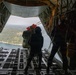The image is taken from inside a military cargo aircraft, likely a C-130 Hercules, with the rear ramp open during flight. Two personnel in dark clothing are visible near the open ramp, with one appearing to hold or push a cargo bundle equipped with red deployment parachute components. Below, a forested landscape with a body of water is visible, suggesting a low-to-medium altitude airdrop operation.