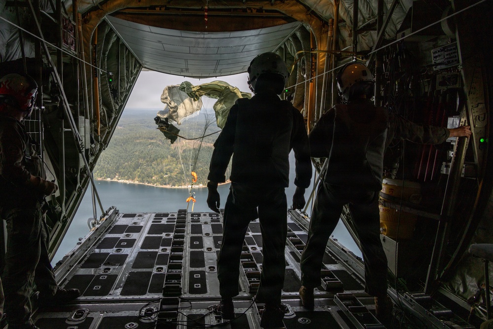 The image shows the interior of a military cargo aircraft, likely a C-130 Hercules, with its rear ramp open during flight. Personnel in flight suits and helmets are observing or supervising what appears to be an airdrop operation, with a deployed parachute and suspended cargo visible outside the aircraft. Below, a forested landscape with a body of water is visible.