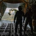 The image shows the interior of a military cargo aircraft, likely a C-130 Hercules, with its rear ramp open during flight. Personnel in flight suits and helmets are observing or supervising what appears to be an airdrop operation, with a deployed parachute and suspended cargo visible outside the aircraft. Below, a forested landscape with a body of water is visible.