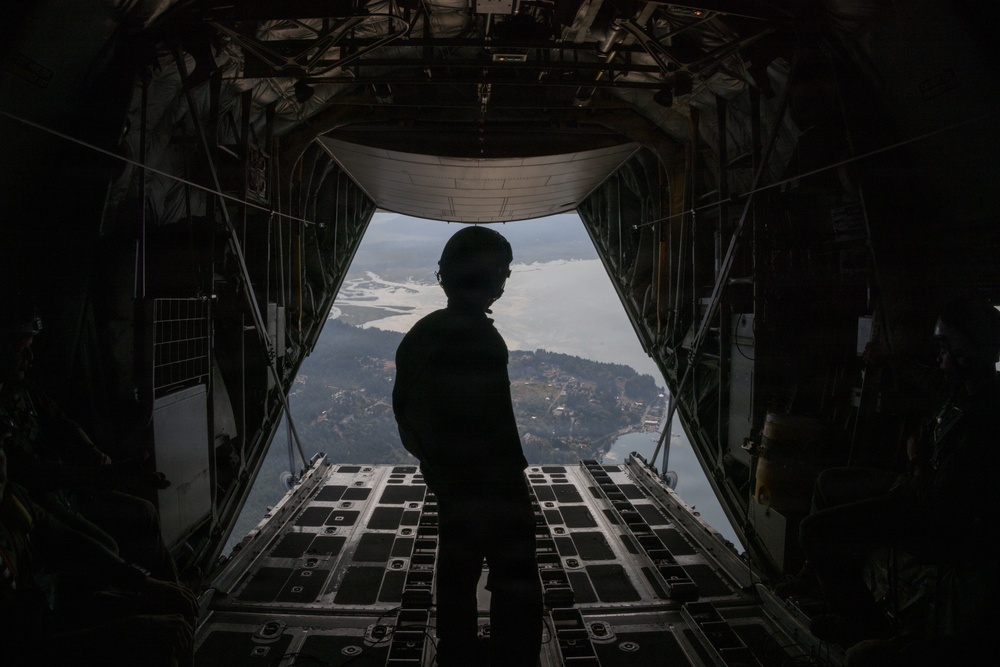 A silhouetted figure wearing a flight helmet stands at the open rear cargo ramp of a military transport aircraft in flight. The open ramp reveals an aerial view of a coastal or lakeside landscape below, with visible terrain, water bodies, and vegetation. The interior of the aircraft cargo bay is visible in the foreground and sides of the frame.