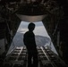 A silhouetted figure wearing a flight helmet stands at the open rear cargo ramp of a military transport aircraft in flight. The open ramp reveals an aerial view of a coastal or lakeside landscape below, with visible terrain, water bodies, and vegetation. The interior of the aircraft cargo bay is visible in the foreground and sides of the frame.