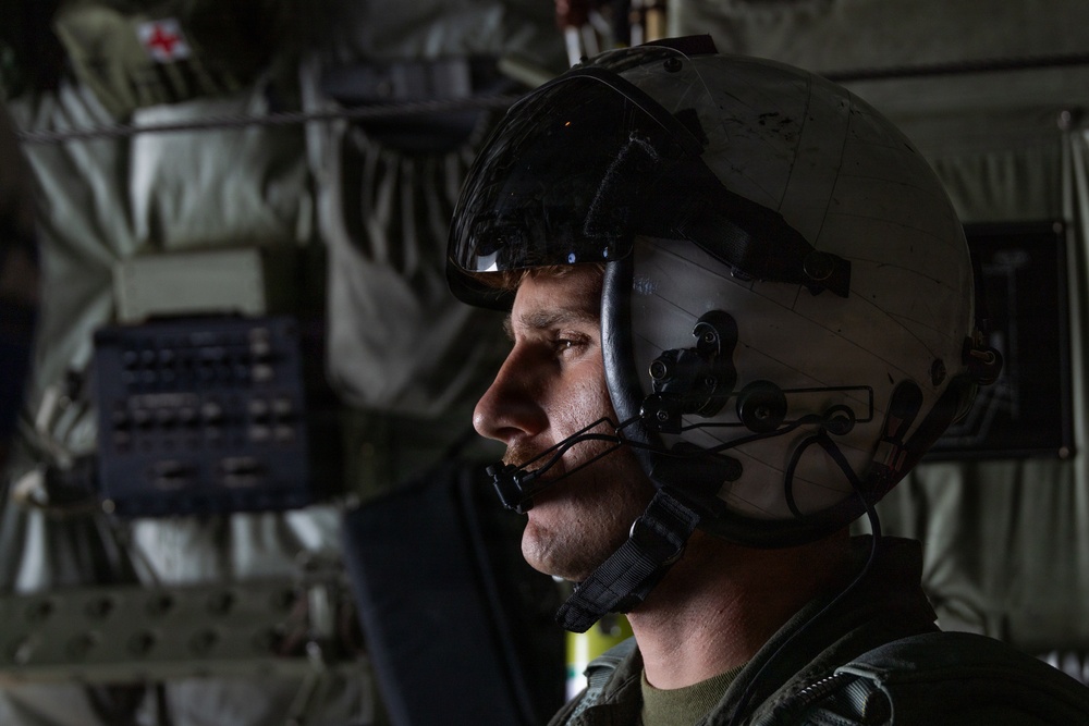 A military aviator or aircrew member is photographed in profile inside what appears to be a military aircraft or helicopter. The subject is wearing a flight helmet with a visor and boom microphone, suggesting active flight or mission operations. The background reveals the interior of a military aircraft with olive-drab equipment, strapping, and what appears to be a medical cross symbol.