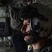 A military aviator or aircrew member is photographed in profile inside what appears to be a military aircraft or helicopter. The subject is wearing a flight helmet with a visor and boom microphone, suggesting active flight or mission operations. The background reveals the interior of a military aircraft with olive-drab equipment, strapping, and what appears to be a medical cross symbol.