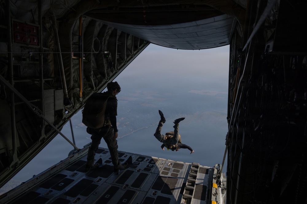 A military parachute operation is captured from inside the open rear cargo ramp of a large military transport aircraft, likely a C-130 Hercules. One person has already jumped and is visible in freefall outside the aircraft, while a second person in tactical gear stands on the ramp observing or preparing to jump. The terrain and water below are visible at high altitude.