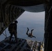 A military parachute operation is captured from inside the open rear cargo ramp of a large military transport aircraft, likely a C-130 Hercules. One person has already jumped and is visible in freefall outside the aircraft, while a second person in tactical gear stands on the ramp observing or preparing to jump. The terrain and water below are visible at high altitude.