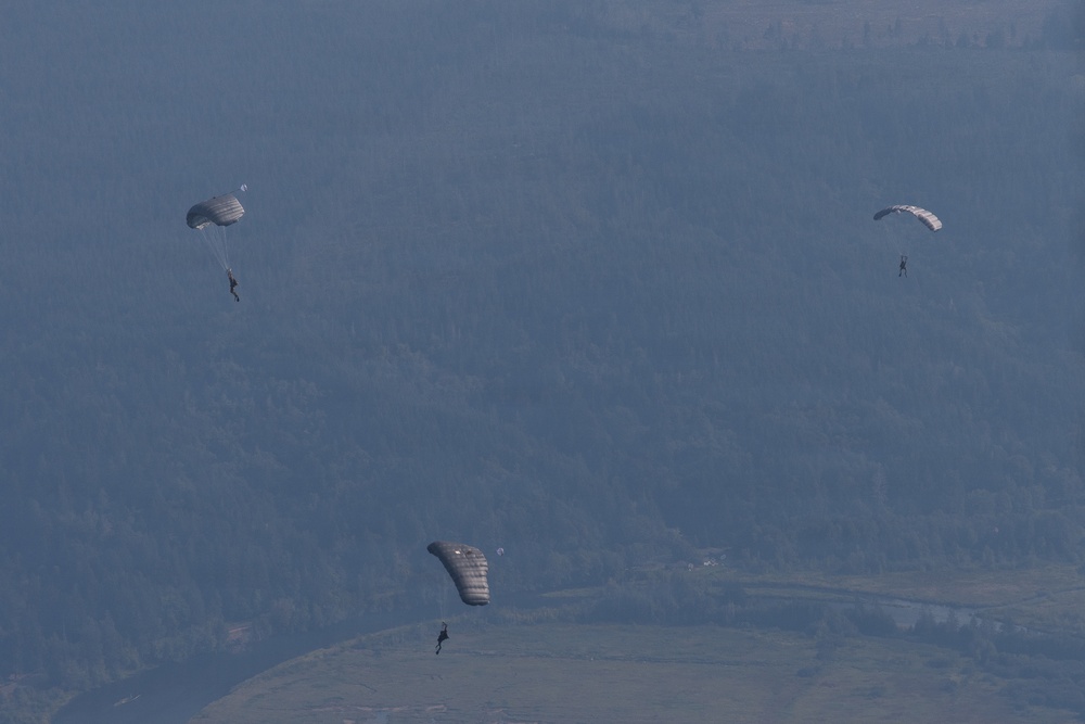 Three parachutists are descending over a mountainous, forested landscape. The parachutes appear to be military-style ram-air canopies in gray/olive tones. The image is taken from an elevated vantage point, possibly from an aircraft, showing the jumpers at varying altitudes above a valley.