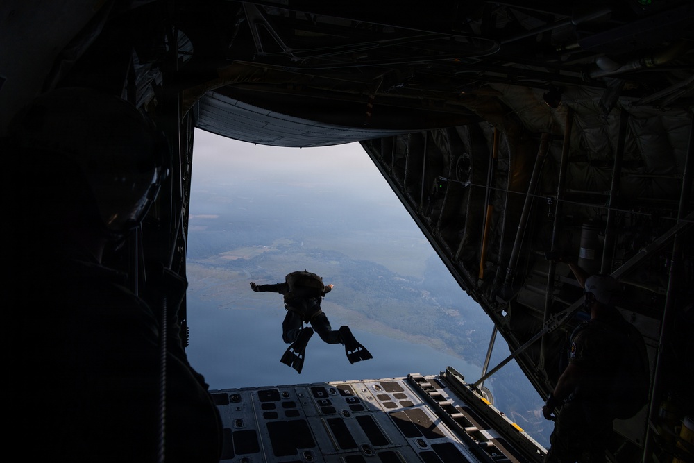 A person in full tactical gear, including fins on their feet, is captured mid-jump from the open rear cargo ramp of a large military transport aircraft. The jumper appears to be performing a HALO (High Altitude Low Opening) or combat dive insertion, evidenced by the diving fins and equipment pack. The landscape below shows a coastal or lakeside terrain visible through the open ramp.