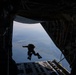 A person in full tactical gear, including fins on their feet, is captured mid-jump from the open rear cargo ramp of a large military transport aircraft. The jumper appears to be performing a HALO (High Altitude Low Opening) or combat dive insertion, evidenced by the diving fins and equipment pack. The landscape below shows a coastal or lakeside terrain visible through the open ramp.