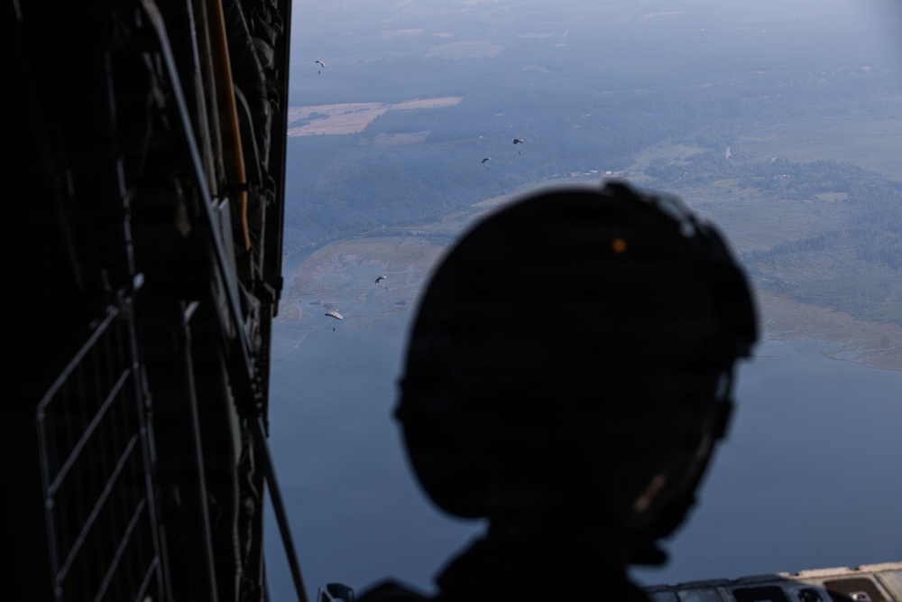 A military personnel member wearing a tactical helmet is silhouetted in the foreground, observing from the open rear ramp of a military transport aircraft. Multiple parachutists with deployed parachutes are visible descending toward a landscape below that includes a body of water, fields, and forested terrain. The image captures an active airborne operation from the perspective of someone inside the aircraft.