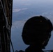 A military personnel member wearing a tactical helmet is silhouetted in the foreground, observing from the open rear ramp of a military transport aircraft. Multiple parachutists with deployed parachutes are visible descending toward a landscape below that includes a body of water, fields, and forested terrain. The image captures an active airborne operation from the perspective of someone inside the aircraft.