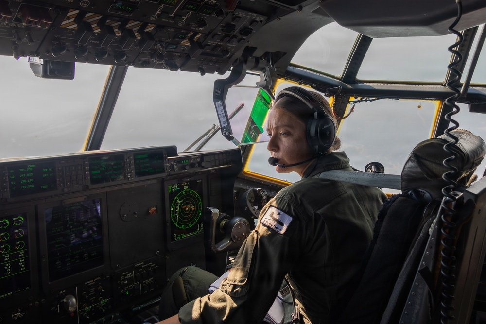 A military pilot, appearing to be female, is seated in the cockpit of a large military aircraft, likely a C-130 Hercules or similar transport aircraft. She is wearing an olive green flight suit and a headset with a boom microphone, actively monitoring instruments and looking out the forward windows. The cockpit displays numerous green-lit avionics panels, navigation displays, and overhead control panels consistent with a modern military transport aircraft.