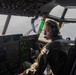 A military pilot, appearing to be female, is seated in the cockpit of a large military aircraft, likely a C-130 Hercules or similar transport aircraft. She is wearing an olive green flight suit and a headset with a boom microphone, actively monitoring instruments and looking out the forward windows. The cockpit displays numerous green-lit avionics panels, navigation displays, and overhead control panels consistent with a modern military transport aircraft.