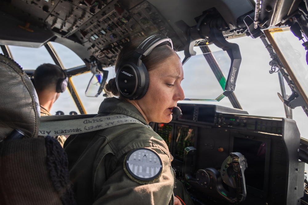 A female military aviator is seated in the cockpit of a large military aircraft, wearing a green flight suit and Bose aviation headset with a boom microphone. She appears to be actively communicating or monitoring instruments during flight. A second crew member is visible in the background at another crew station.