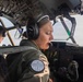 A female military aviator is seated in the cockpit of a large military aircraft, wearing a green flight suit and Bose aviation headset with a boom microphone. She appears to be actively communicating or monitoring instruments during flight. A second crew member is visible in the background at another crew station.