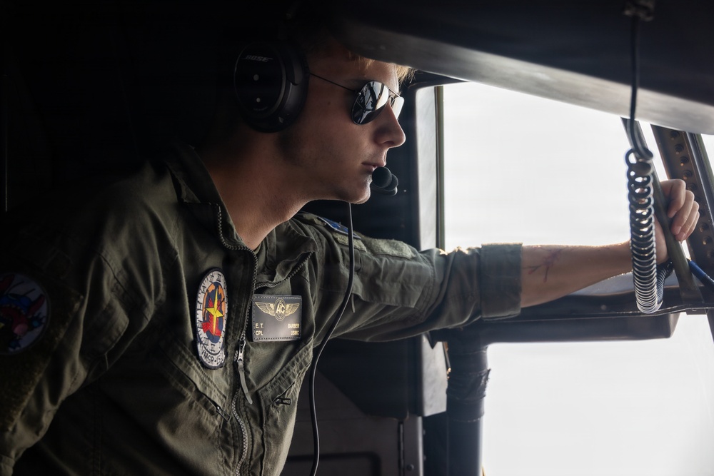 A military aviator wearing an olive green flight suit is seated in an aircraft cockpit, looking out through an open window or hatch. The individual is equipped with a Bose aviation headset, a boom microphone, and reflective aviator sunglasses. Patches on the flight suit suggest affiliation with a Marine Corps aviation unit (VMGR designation visible).