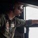 A military aviator wearing an olive green flight suit is seated in an aircraft cockpit, looking out through an open window or hatch. The individual is equipped with a Bose aviation headset, a boom microphone, and reflective aviator sunglasses. Patches on the flight suit suggest affiliation with a Marine Corps aviation unit (VMGR designation visible).