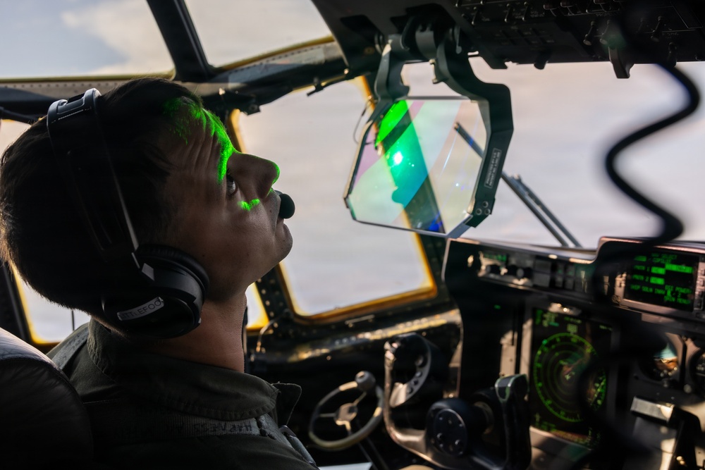 A military pilot or aircrew member is seated in a large aircraft cockpit, illuminated by a distinctive green laser or light source reflecting off their face and a heads-up display (HUD) panel. The cockpit features multiple analog and digital instruments, suggesting a large military transport or patrol aircraft. The green light appears to be emanating from or reflecting through a HUD or optical device mounted near the windscreen.