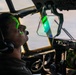 A military pilot or aircrew member is seated in a large aircraft cockpit, illuminated by a distinctive green laser or light source reflecting off their face and a heads-up display (HUD) panel. The cockpit features multiple analog and digital instruments, suggesting a large military transport or patrol aircraft. The green light appears to be emanating from or reflecting through a HUD or optical device mounted near the windscreen.