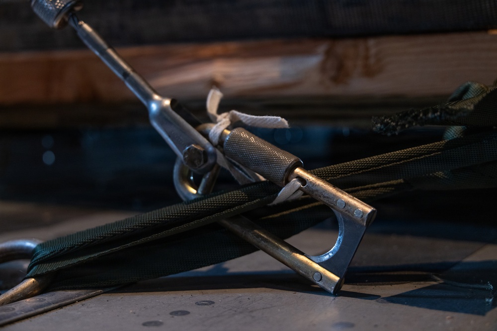 A close-up photograph of metal rigging or tensioning hardware, likely turnbuckles or similar fastening devices, resting on a flat surface alongside green nylon webbing straps and rope. The metal components show signs of wear and use, with knurled grips and clevis-style end fittings visible. The image appears to depict equipment used in sailing, rigging, or load-securing applications.