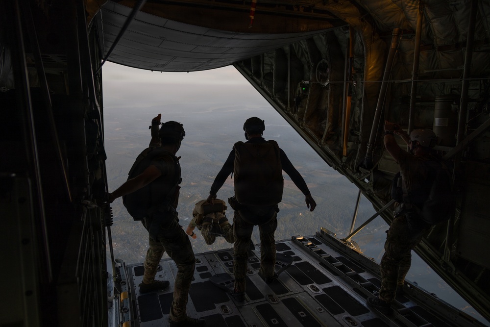 Military personnel are standing at the open rear cargo ramp of a large military transport aircraft, appearing to prepare for a parachute jump. Two figures are silhouetted against the open sky at the edge of the ramp, with terrain visible far below. A third individual is visible on the right side, possibly a jumpmaster or crew member.
