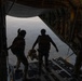Military personnel are standing at the open rear cargo ramp of a large military transport aircraft, appearing to prepare for a parachute jump. Two figures are silhouetted against the open sky at the edge of the ramp, with terrain visible far below. A third individual is visible on the right side, possibly a jumpmaster or crew member.