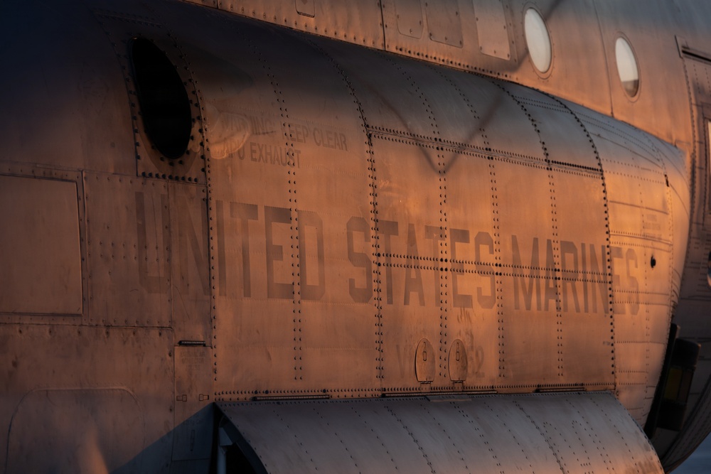 Close-up photograph of the fuselage of a United States Marines military aircraft, illuminated by warm golden-hour light. The metal skin shows riveted panel construction typical of large military transport or helicopter aircraft. The image captures stenciled text, circular windows (portholes), and maintenance markings on the aircraft's exterior.