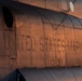 Close-up photograph of the fuselage of a United States Marines military aircraft, illuminated by warm golden-hour light. The metal skin shows riveted panel construction typical of large military transport or helicopter aircraft. The image captures stenciled text, circular windows (portholes), and maintenance markings on the aircraft's exterior.