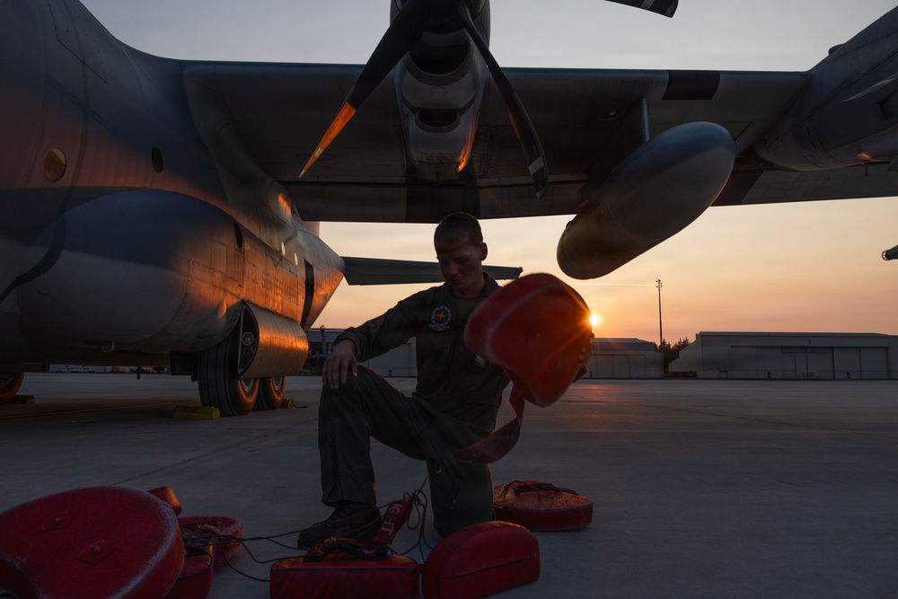 A military aviation crew member in a flight suit performs pre-flight or post-flight ground checks beneath a large military tiltrotor aircraft, likely a V-22 Osprey, during sunrise or sunset. The technician is handling red safety covers or chocks on the flight line of what appears to be a military air base. The warm golden light of the low sun creates dramatic backlighting across the tarmac and aircraft.