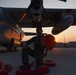 A military aviation crew member in a flight suit performs pre-flight or post-flight ground checks beneath a large military tiltrotor aircraft, likely a V-22 Osprey, during sunrise or sunset. The technician is handling red safety covers or chocks on the flight line of what appears to be a military air base. The warm golden light of the low sun creates dramatic backlighting across the tarmac and aircraft.
