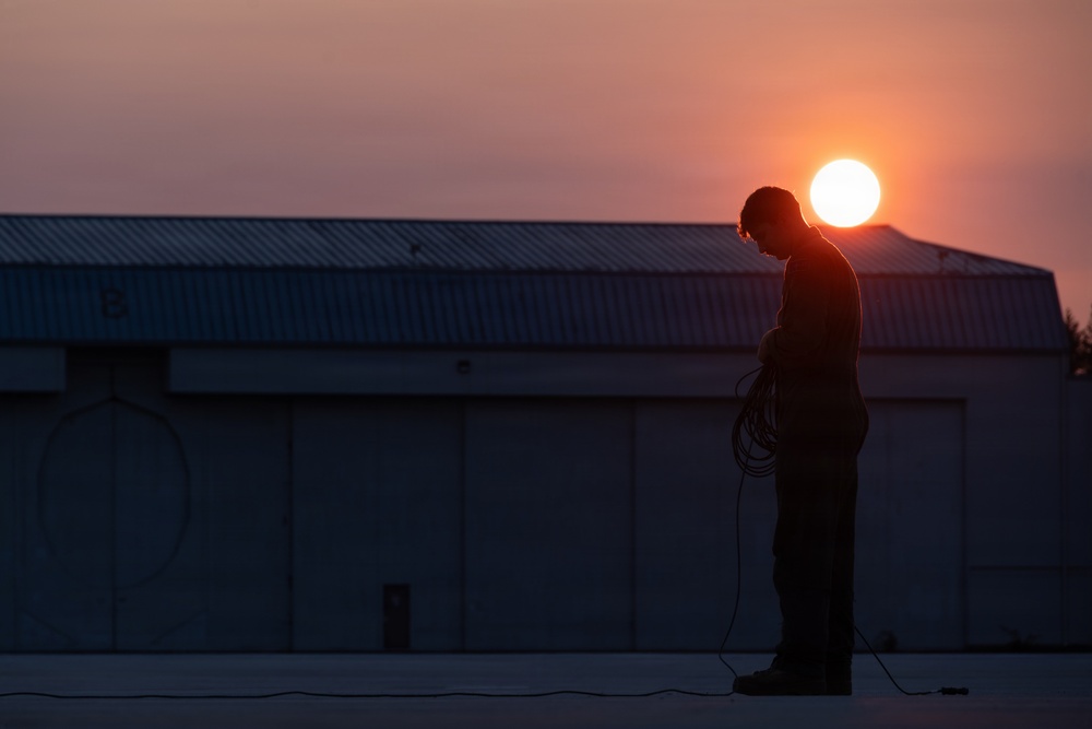 A silhouetted figure stands on an elevated flat surface, head bowed, holding a coiled cable or rope. The scene is backlit by a vivid sunset with a bright solar disk positioned just above the figure's head. A large industrial or hangar-style building with a corrugated metal roof is visible in the background.